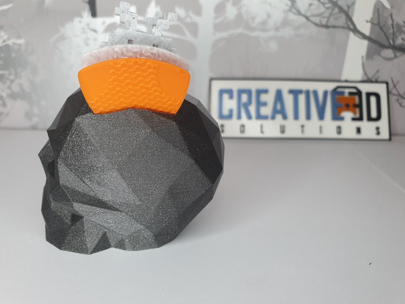 Puede incluir: Un cr&aacute;neo negro impreso en 3D con un dise&ntilde;o geom&eacute;trico. Una peque&ntilde;a nave espacial impresa en 3D de color naranja y blanco se encuentra en la parte superior del cr&aacute;neo. El fondo es una imagen borrosa de una rama de &aacute;rbol y un letrero que dice "CREATIVE 3D SOLUTIONS".