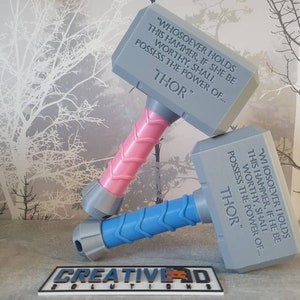 May include: Two gray 3D printed replicas of Thor's hammer, Mjolnir, with the inscription "Whosoever holds this hammer, if she be worthy, shall possess the power of... Thor." One hammer has a pink handle and the other has a blue handle.