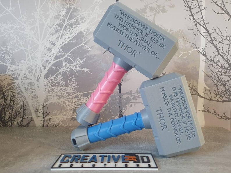 May include: Two gray 3D printed replicas of Thor's hammer, Mjolnir, with the inscription "Whosoever holds this hammer, if she be worthy, shall possess the power of... Thor." One hammer has a pink handle and the other has a blue handle.