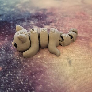 May include: A grey 3D printed cat figurine with a segmented body, lying on a purple and pink galaxy-themed surface.