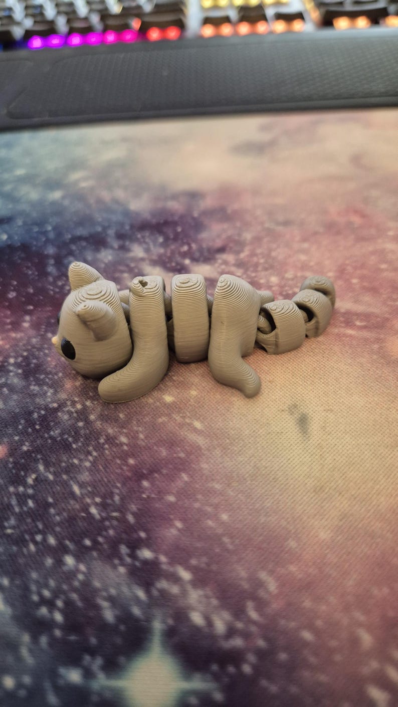May include: A grey 3D printed cat figurine with a segmented body, lying on a purple and pink galaxy-themed surface.