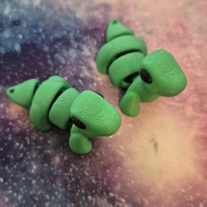 May include: Two green articulated dinosaur toys with black eyes. The dinosaurs are posed on a purple and blue galaxy patterned surface.