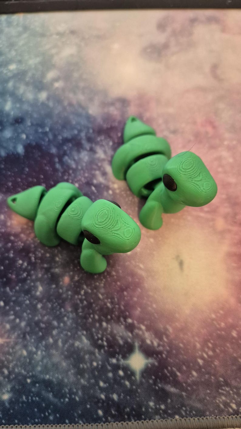 May include: Two green articulated dinosaur toys with black eyes. The dinosaurs are posed on a purple and blue galaxy patterned surface.