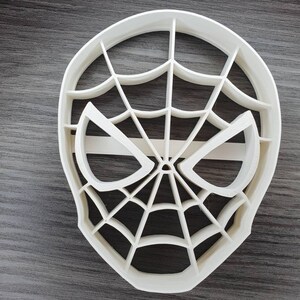 May include: White plastic cookie cutter shaped like a Spiderman mask with a web design.