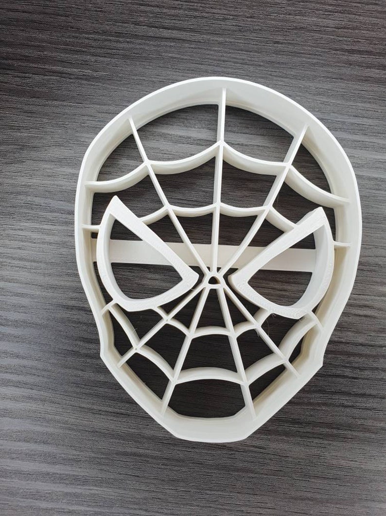 May include: White plastic cookie cutter shaped like a Spiderman mask with a web design.