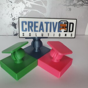 May include: Three 3D printed phone holders in green, blue and pink. Each holder has a square base and a round ball on top. The ball is attached to a small, flat, square piece that is attached to the base. The holders are arranged in a triangle shape. The text "CREATIVE3D SOLUTIONS" is visible in the background.