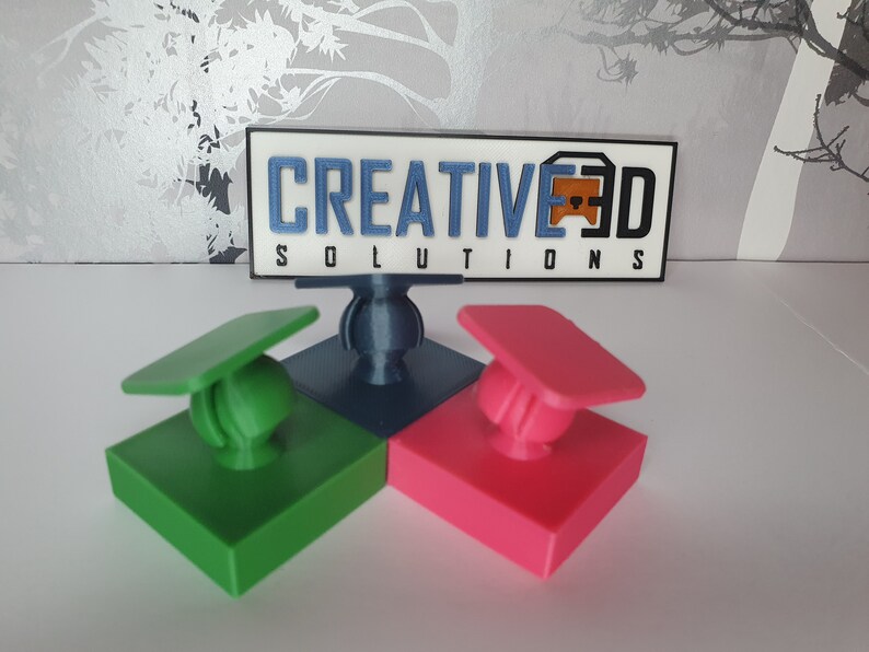 May include: Three 3D printed phone holders in green, blue and pink. Each holder has a square base and a round ball on top. The ball is attached to a small, flat, square piece that is attached to the base. The holders are arranged in a triangle shape. The text "CREATIVE3D SOLUTIONS" is visible in the background.