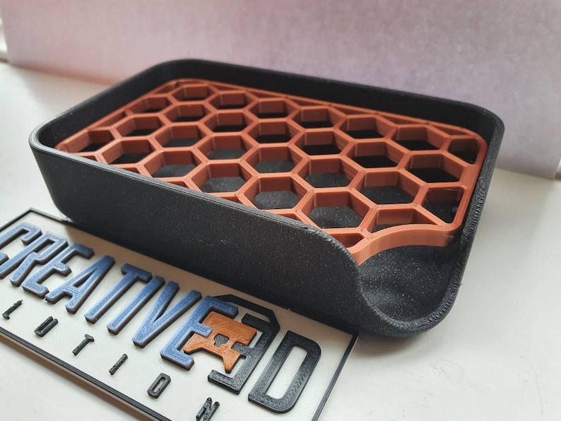 May include: A black soap dish with a honeycomb-shaped insert made of brown plastic. The dish has a curved edge and is designed to drain water.