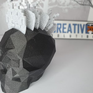 Puede incluir: Un cr&aacute;neo negro geom&eacute;trico con un mohawk blanco y pixelado. El cr&aacute;neo est&aacute; hecho de un material impreso en 3D y tiene una superficie texturizada.