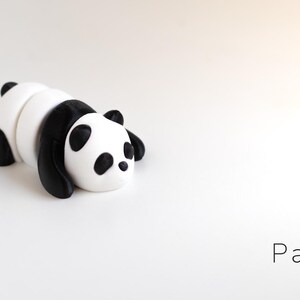 May include: A small, stylized panda figurine in black and white, lying down on a white surface. The panda has black ears, eyes, and limbs, with a white body. The word "Panda" and "Baby" are written in a minimalist font.