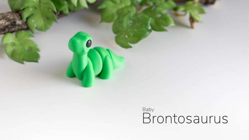 May include: A small, green, plastic dinosaur toy shaped like a baby Brontosaurus. The toy has black eyes and a friendly expression.