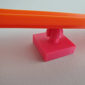 May include: A bright orange plastic track piece is balanced on a pink plastic base with a round knob. The base is square shaped.