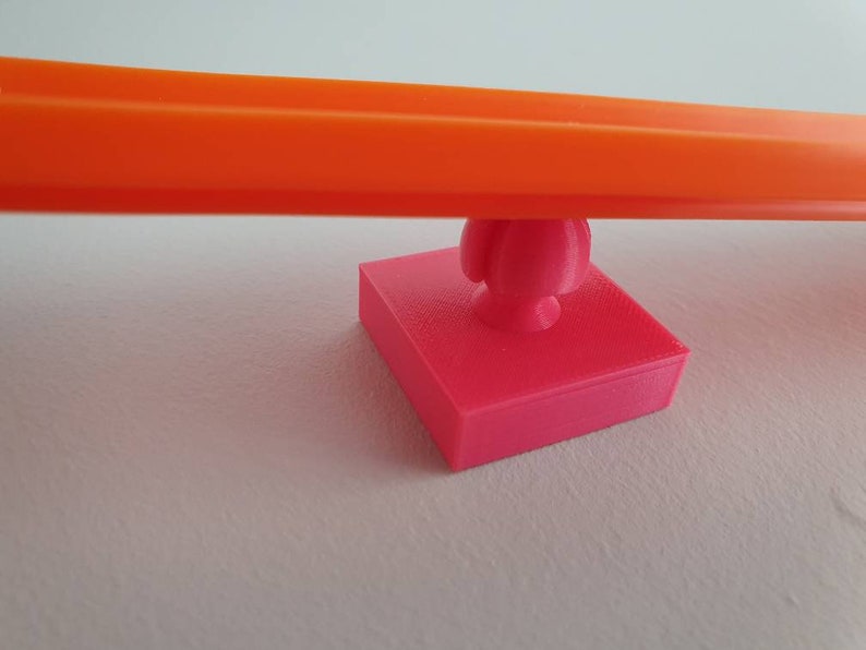 May include: A bright orange plastic track piece is balanced on a pink plastic base with a round knob. The base is square shaped.