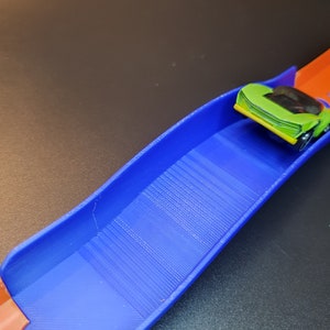 Op de afbeelding: Een blauwe en oranje plastic speelgoed racebaan met een groene speelgoedauto op de baan.