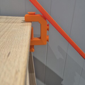 Op de afbeelding: Een oranje plastic klem bevestigd aan een houten oppervlak, die een fel oranje plastic baanstuk vasthoudt.