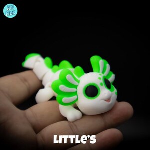 May include: A white and green axolotl figurine with black eyes. The figurine is sitting on a hand.