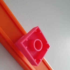 May include: A pink plastic track piece with a round hole in the centre. The track piece is designed to connect to other track pieces to create a race track.
