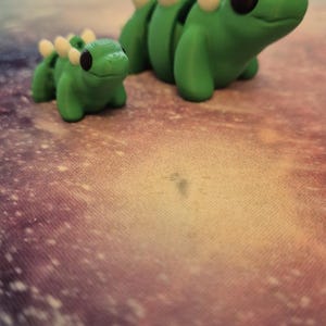 May include: Two green plastic dinosaur figurines with white spines. The larger dinosaur is facing right and the smaller dinosaur is facing left.