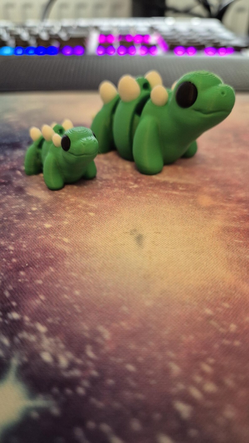 May include: Two green plastic dinosaur figurines with white spines. The larger dinosaur is facing right and the smaller dinosaur is facing left.