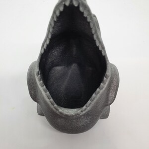 May include: A black, 3D printed dinosaur planter with an open mouth. The planter is made of a textured material and has a speckled finish.