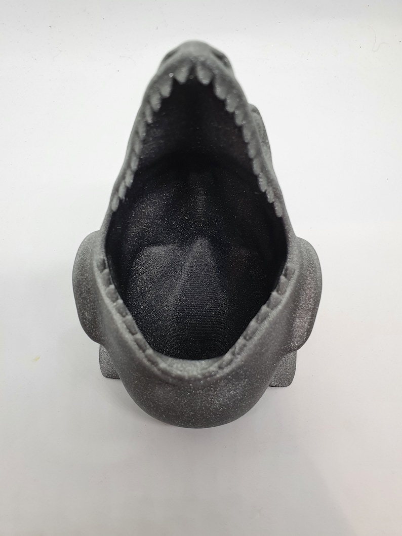 May include: A black, 3D printed dinosaur planter with an open mouth. The planter is made of a textured material and has a speckled finish.