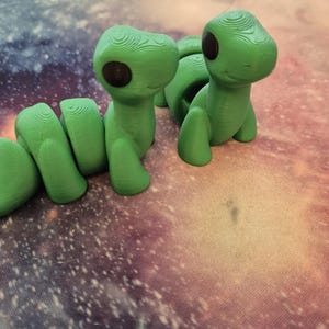 May include: Two green 3D printed figurines of a worm with black eyes. The worm on the left is curled up and the worm on the right is standing upright.