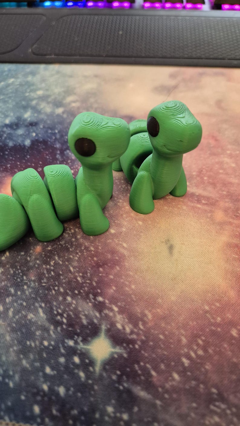 May include: Two green 3D printed figurines of a worm with black eyes. The worm on the left is curled up and the worm on the right is standing upright.