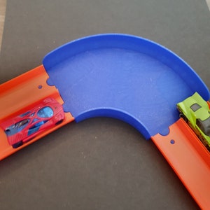 May include: A blue plastic track piece for a toy car race track. The track piece is curved and has two orange track pieces connected to it. A red toy car is on the left track and a green toy car is on the right track.