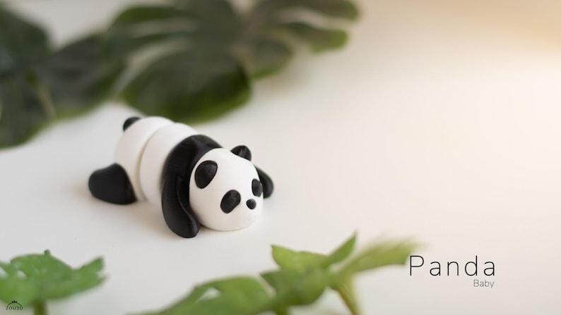May include: A handmade panda figurine in black and white, lying down on a white surface. The panda has black ears, eyes, and limbs. The word "Panda Baby" is written in a minimalist font.