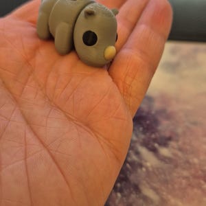 May include: A small, gray, 3D printed figurine of a rhinoceros with a light brown nose. The figurine is sitting in the palm of a hand.