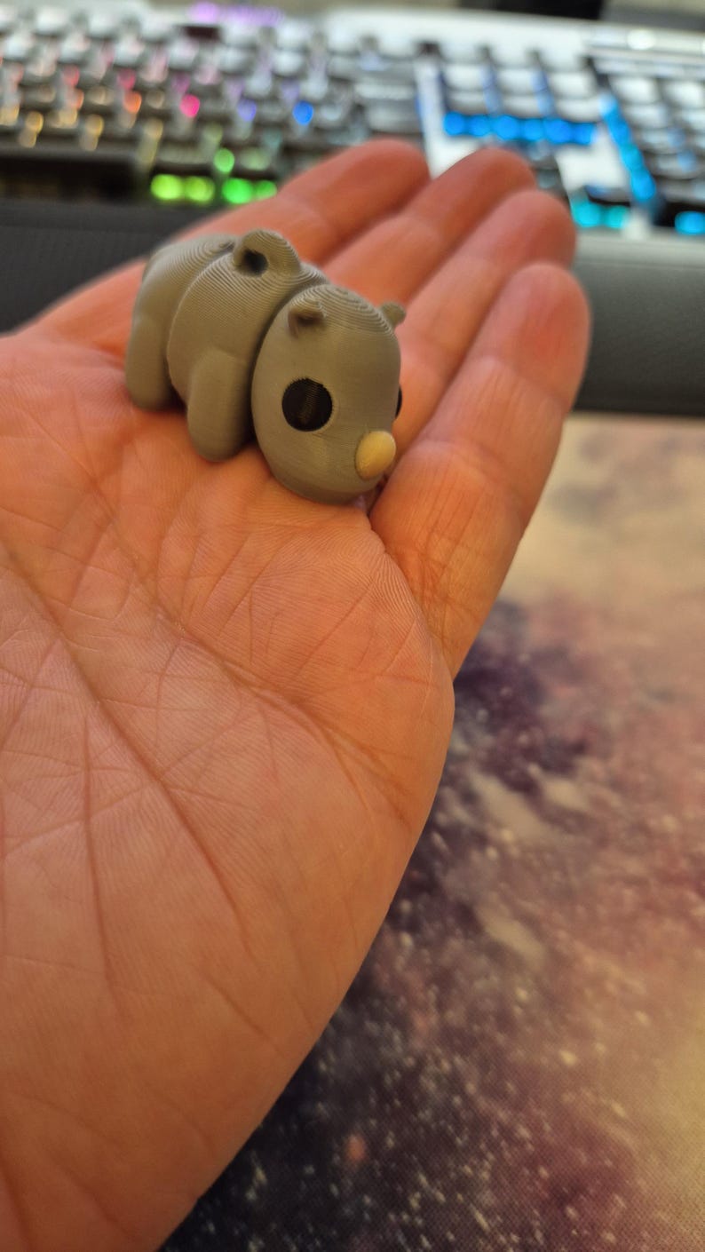 May include: A small, gray, 3D printed figurine of a rhinoceros with a light brown nose. The figurine is sitting in the palm of a hand.