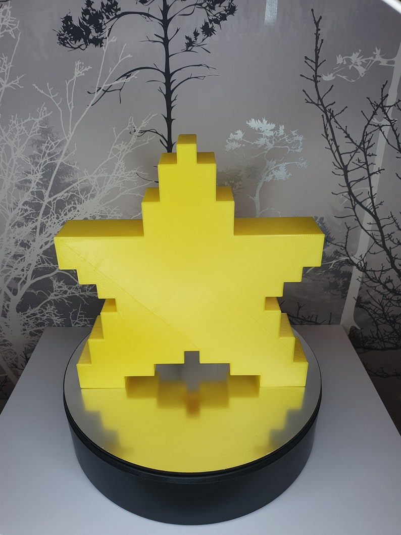 Puede incluir: Una estrella amarilla impresa en 3D sobre una base giratoria negra. La estrella est&aacute; formada por peque&ntilde;os cuadrados y tiene un aspecto pixelado.