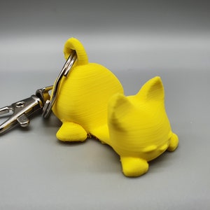 May include: A yellow 3D printed keychain shaped like a dog. The dog is lying down with its head resting on its paws. The keychain has a silver metal ring.