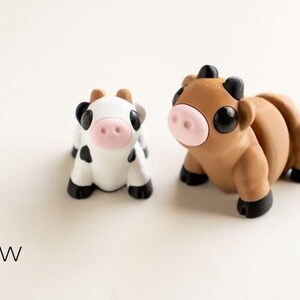May include: Two miniature cow figurines. One is white with black spots, the other is brown. Both have pink noses, black hooves, and black horns. The text "Tiny Cow" is below the figurines.