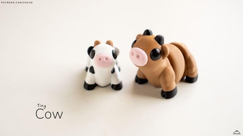 May include: Two miniature cow figurines. One is white with black spots, the other is brown. Both have pink noses, black hooves, and black horns. The text "Tiny Cow" is below the figurines.