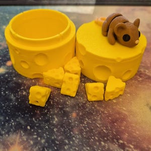 Puede incluir: Un recipiente amarillo con forma de queso con una figurita de rat&oacute;n en la parte superior. El recipiente est&aacute; abierto y hay varias piezas peque&ntilde;as de queso esparcidas a su alrededor.