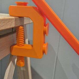 May include: Orange plastic clamp with a screw and two rubber feet. The clamp is attached to a wooden table.