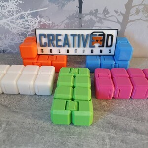 Op de afbeelding: Vijf verschillende fidget-speelgoedjes, elk gemaakt van een ander gekleurd plastic. De speelgoedjes zijn in een rij gerangschikt, met het witte speelgoedje in het midden. De speelgoedjes zijn allemaal gemaakt van zacht, flexibel plastic en hebben verschillende texturen. De speelgoedjes zijn allemaal ongeveer 12,5 cm lang en 5 cm breed. De speelgoedjes zijn allemaal gelabeld met de tekst "CREATIVE3D SOLUTIONS".