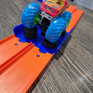 May include: A red and yellow monster truck toy with blue wheels on a bright orange track. The track has a blue plastic ramp.