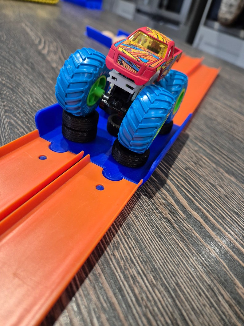 May include: A red and yellow monster truck toy with blue wheels on a bright orange track. The track has a blue plastic ramp.