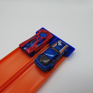 May include: Two toy race cars, one red and blue, the other blue with orange stripes and the number 20, are positioned on a blue starting gate. The cars are on an orange race track.
