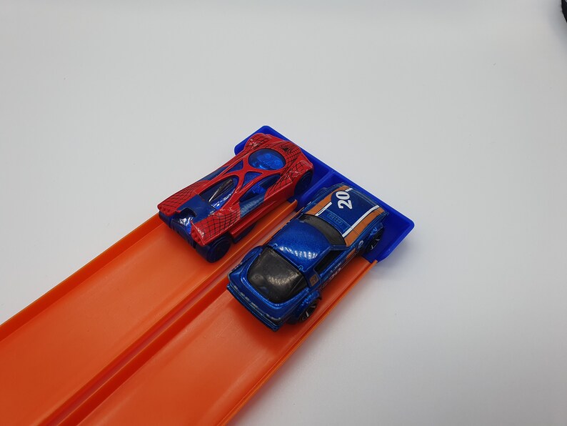 May include: Two toy race cars, one red and blue, the other blue with orange stripes and the number 20, are positioned on a blue starting gate. The cars are on an orange race track.