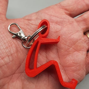 May include: Red 3D printed keychain shaped like a person sitting down. The keychain has a silver split ring and a silver jump ring.