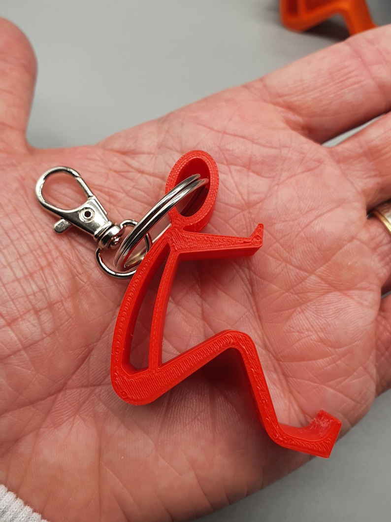 May include: Red 3D printed keychain shaped like a person sitting down. The keychain has a silver split ring and a silver jump ring.