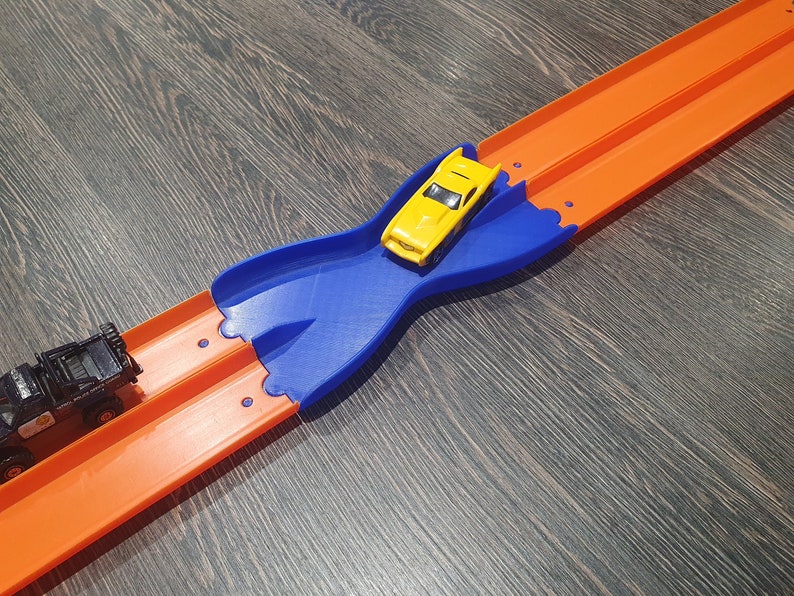 K&ouml;nnte beinhalten: Ein blaues Plastik-Streckenst&uuml;ck f&uuml;r eine Spielzeug-Autorennstrecke. Das Streckenst&uuml;ck ist in Form einer Acht geformt und hat zwei orangefarbene Streckenst&uuml;cke, die daran angeschlossen sind. Ein gelbes Spielzeugauto befindet sich auf dem blauen Streckenst&uuml;ck.