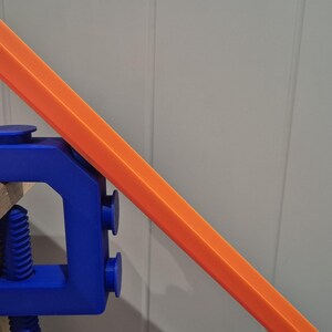 May include: A blue plastic clamp holding a bright orange track piece. The clamp is attached to a wooden surface.