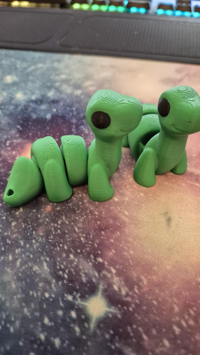 May include: Two green 3D printed worm figurines with black eyes. The worm on the left is curled up and the worm on the right is standing upright.