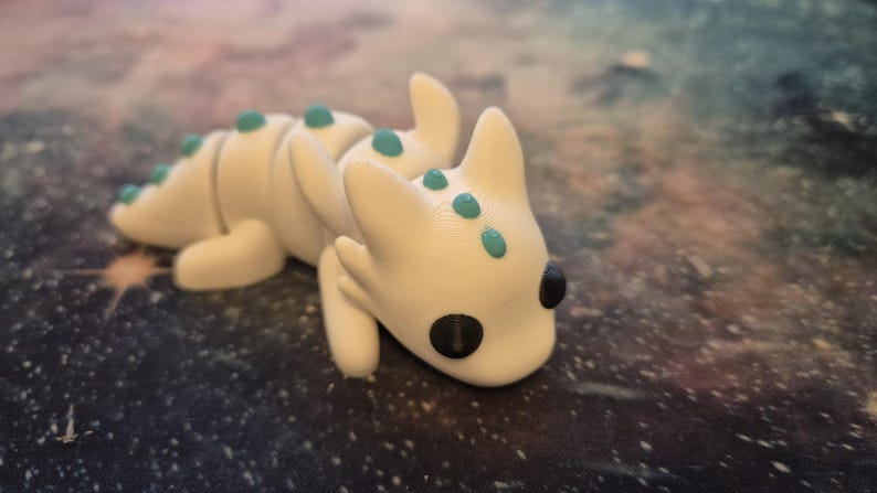 May include: A white dragon figurine with teal accents. The dragon is lying on its belly with its head turned to the side. It has black eyes and a small mouth.