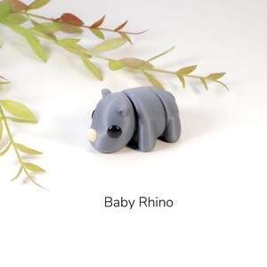 May include: A small gray baby rhino figurine with black eyes and a light brown nose. The figurine is lying on its side with its head turned to the left.