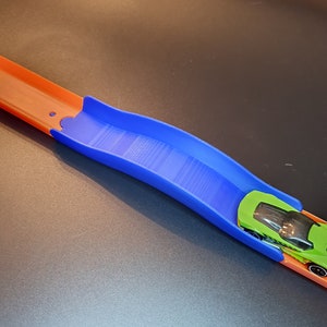 Op de afbeelding: Een blauw plastic baanstuk voor een speelgoedauto racebaan. Het baanstuk is gebogen en een groene speelgoedauto rijdt eroverheen.